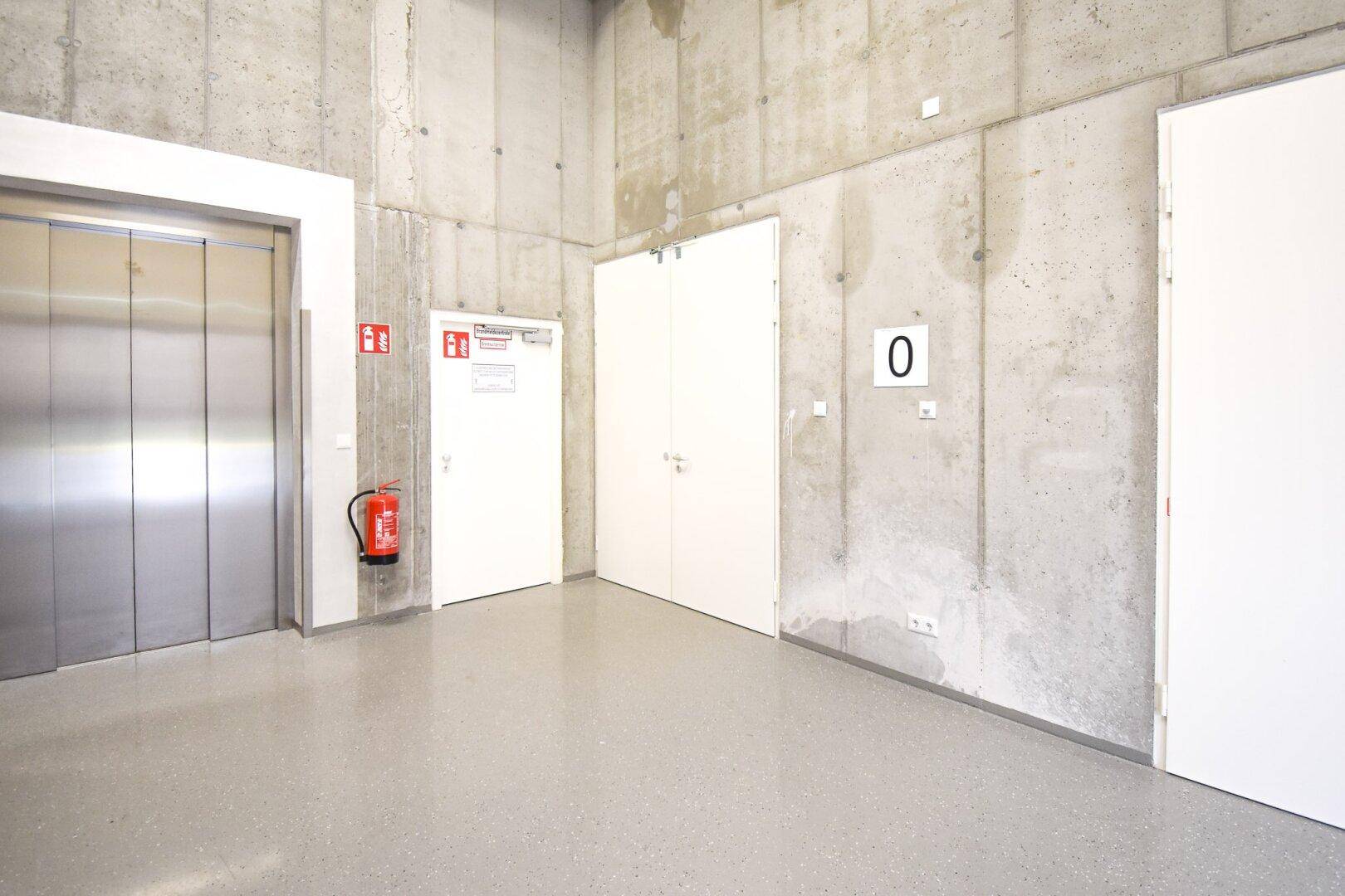 Eingangsbereich mit Aufzug, Sichtbetonwänden, grauem Boden und weißen Türen, gekennzeichnet mit einer "0".