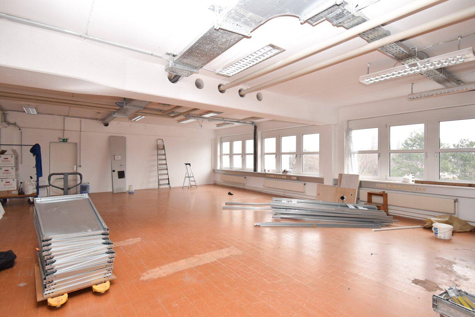 Großer leerer Gewerberaum mit orangefarbenen Fliesen, vielen Fenstern und sichtbaren Installationen an der Decke.