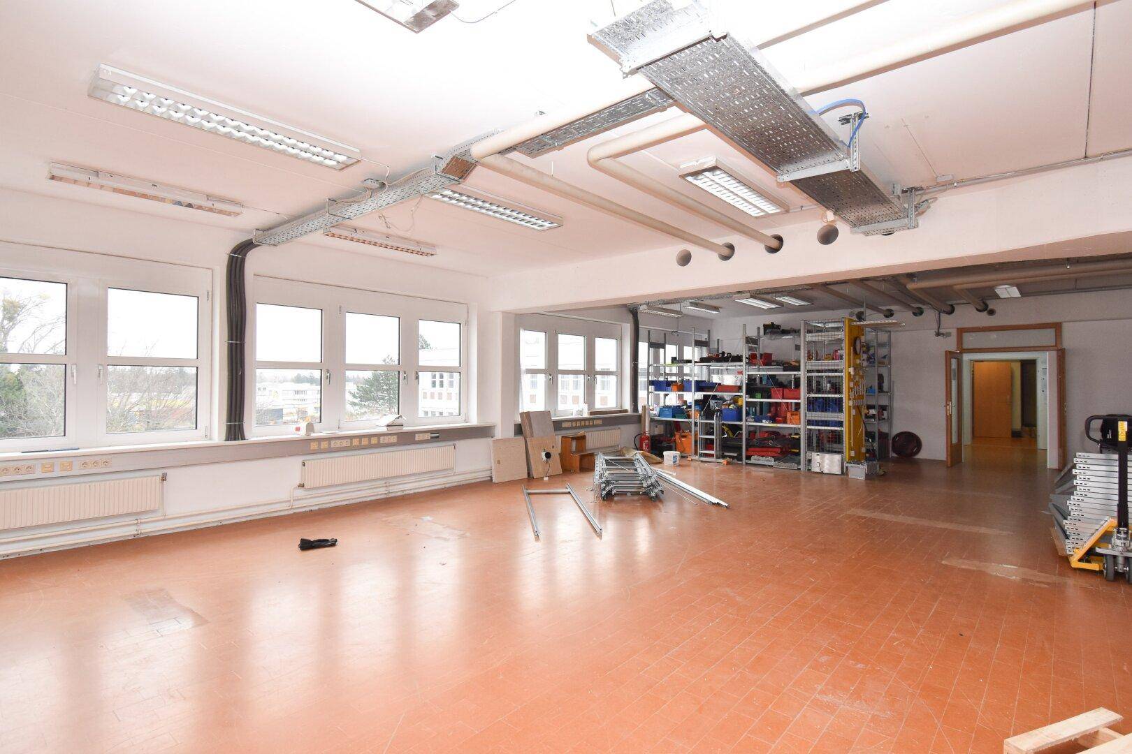 Geräumiger Gewerberaum mit Fliesenboden, großen Fenstern und sichtbaren Deckeninstallationen, teilweise mit Regalen.