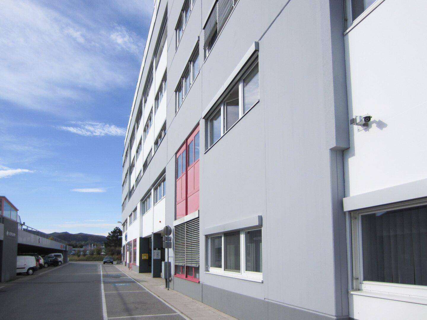 Seitenansicht eines modernen Gebäudes mit grauer Fassade, Rollläden und einer Überwachungskamera unter blauem Himmel.