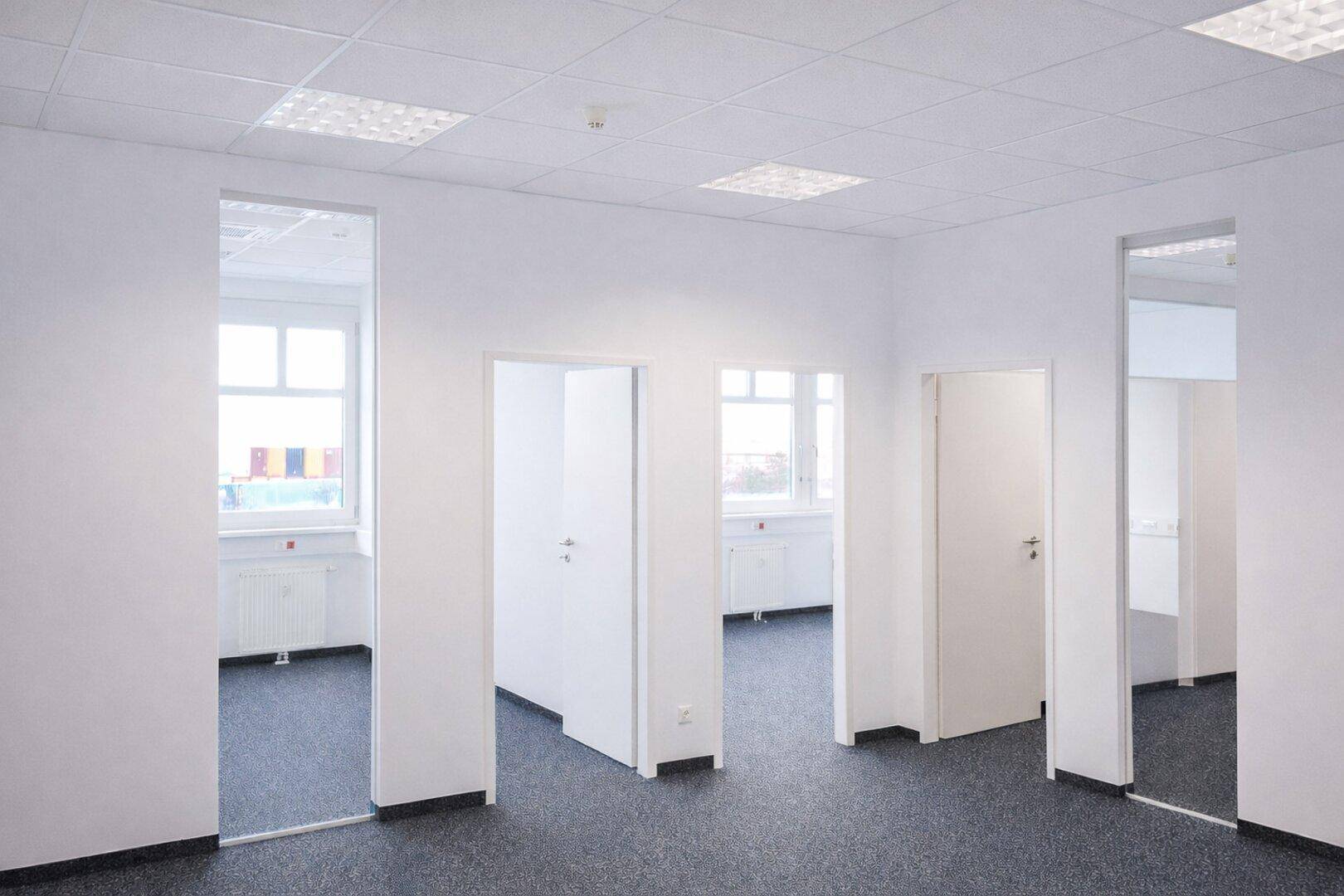 Heller Bürobereich mit mehreren Eingängen zu einzelnen Büros und Teppichboden, ausgestattet mit großen Fenstern.