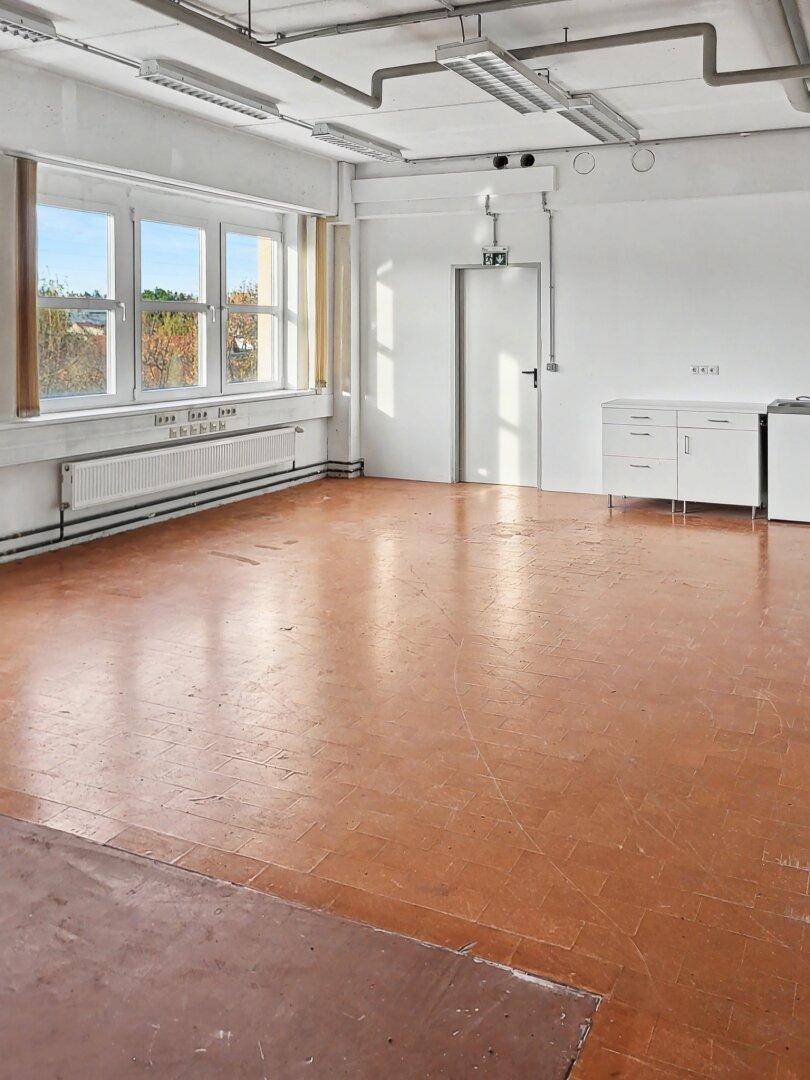 Heller, leerer Gewerberaum mit großen Fenstern, Heizkörpern und sichtbaren Deckeninstallationen.