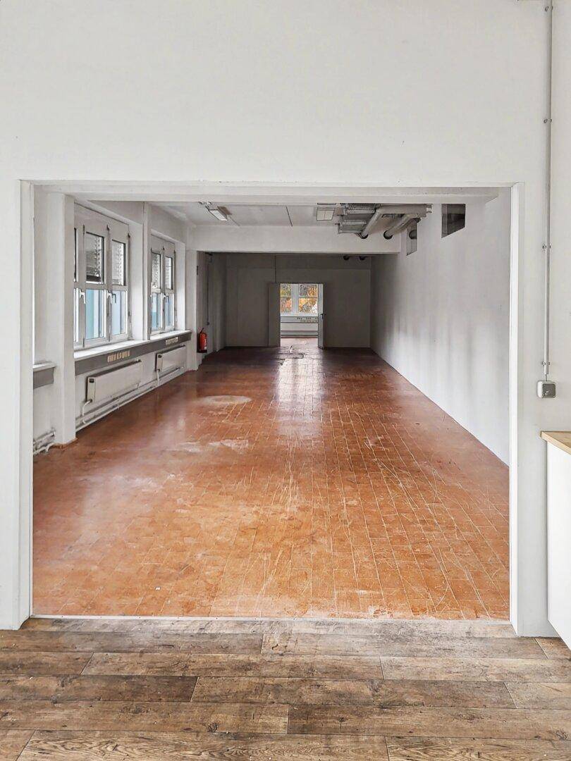 Blick durch eine Tür in einen langen Raum mit Fliesenboden, weißen Wänden und Fenstern.