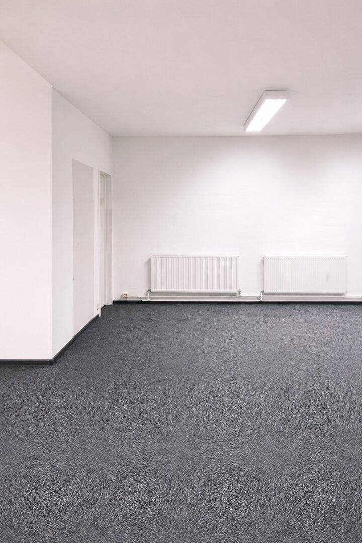 Leerer Büroraum mit grauem Teppichboden, weißen Wänden und Heizkörpern unter der Deckenbeleuchtung.