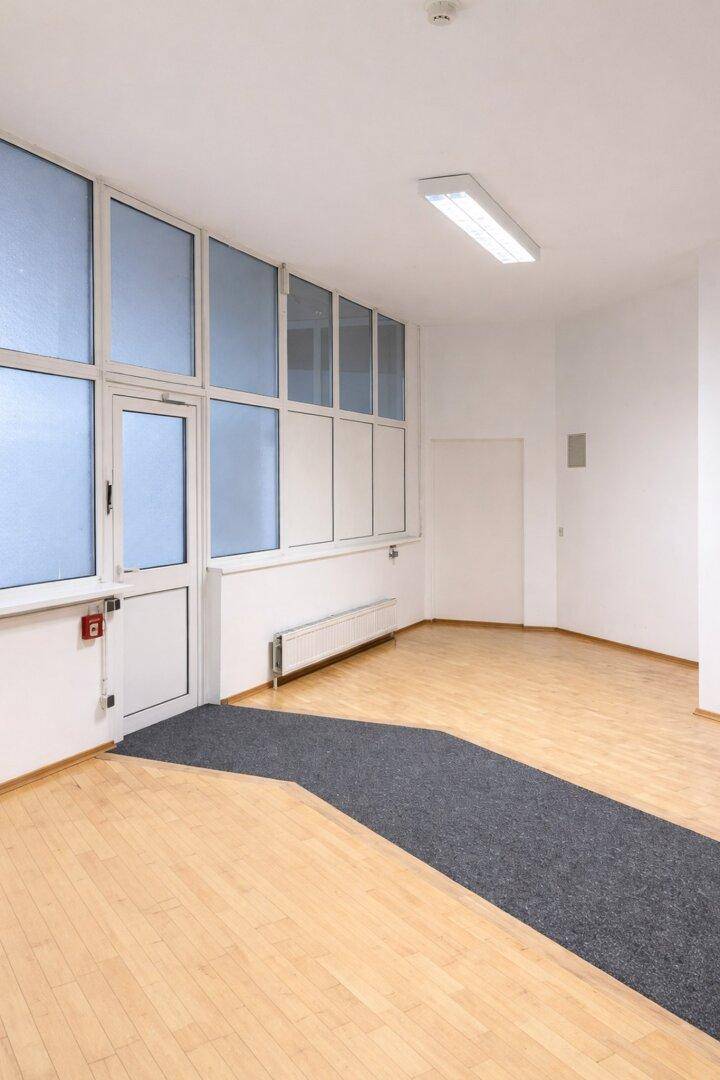 Heller Büroraum mit großen Fenstern, Holzparkettboden und einem Eingangsbereich mit Teppich.