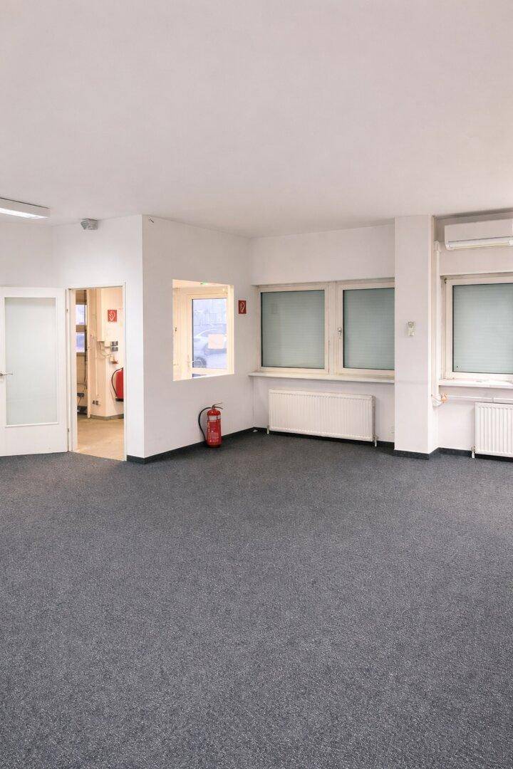 Heller Büroraum mit großen Fenstern, grauem Teppichboden und Heizkörpern, ideal für gewerbliche Nutzung.