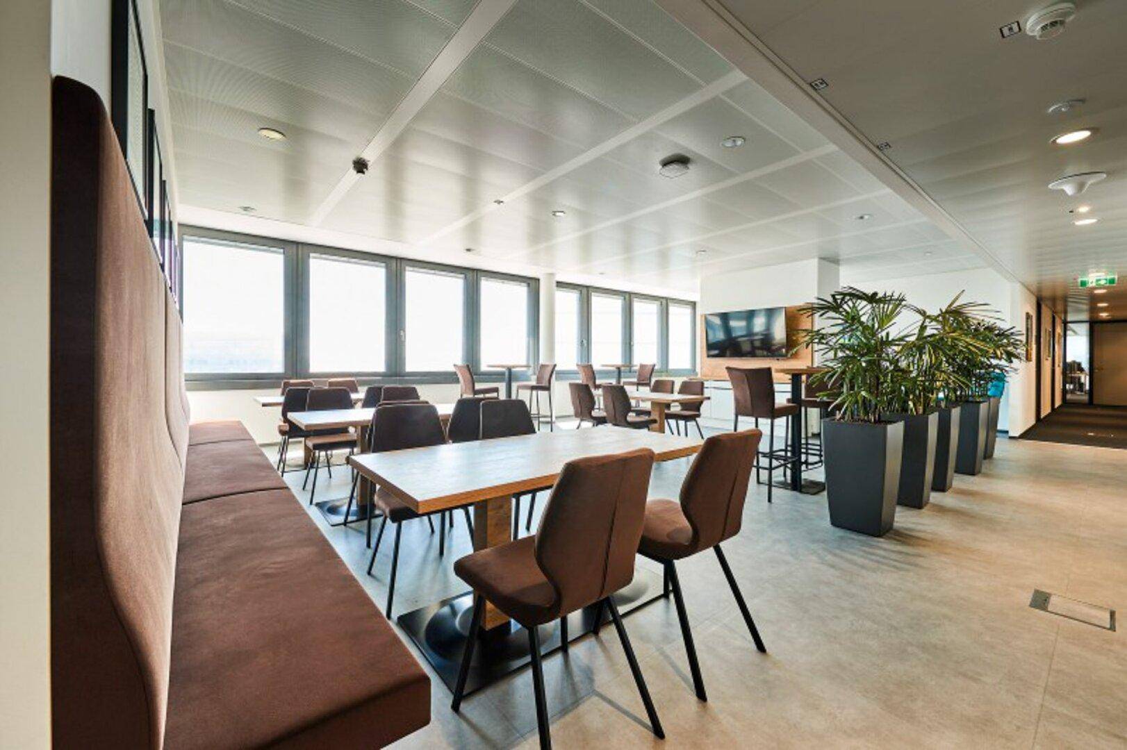 Helle und geräumige Cafeteria mit langen Tischen, bequemen Sitzbänken und großen Fenstern, die eine angenehme Atmosphäre schaffen.