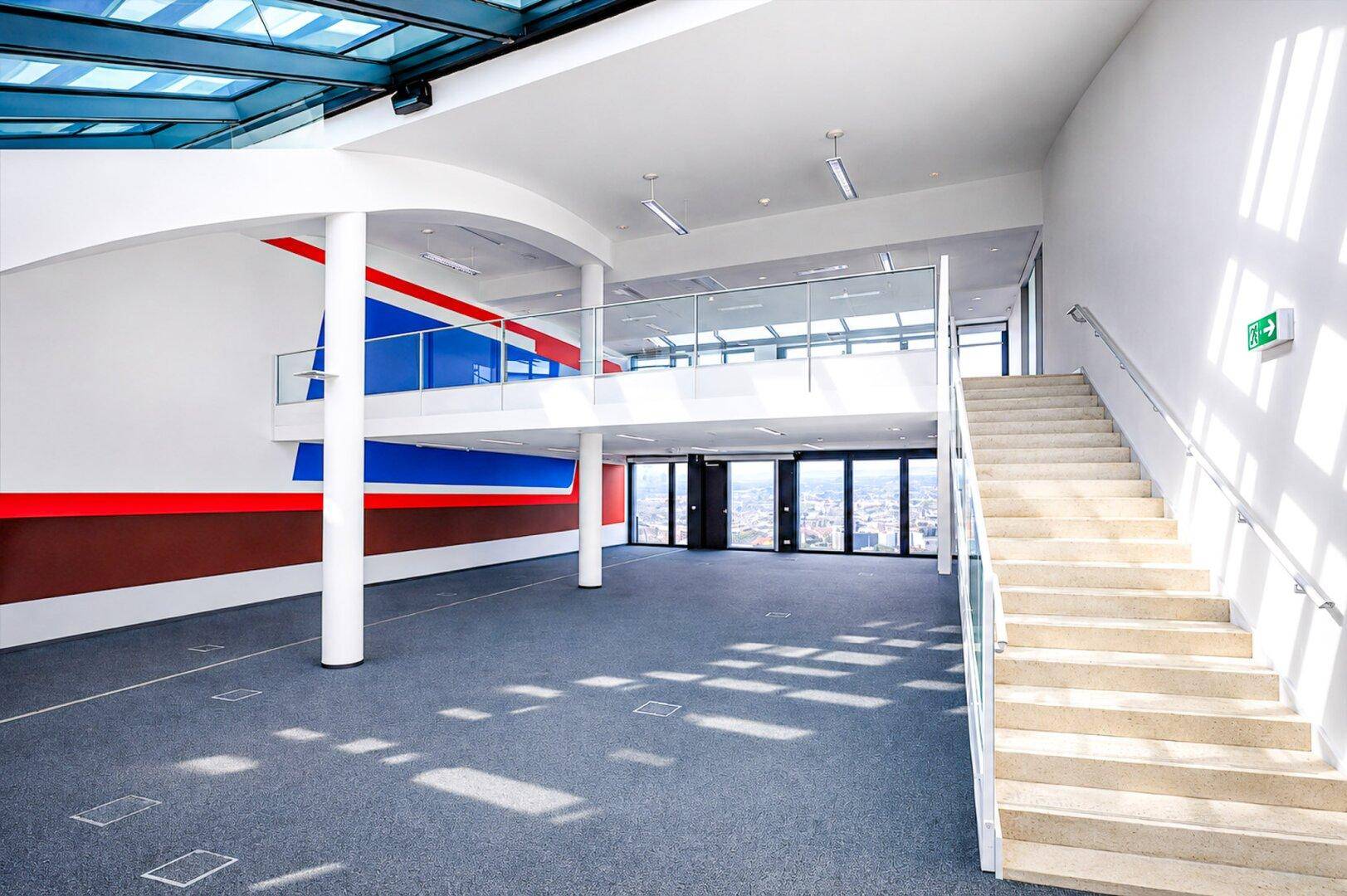 Großer, offener Innenraum mit hohen Decken, einer Galerieebene, breiter Treppe und weitem Stadtblick durch große Fenster.
