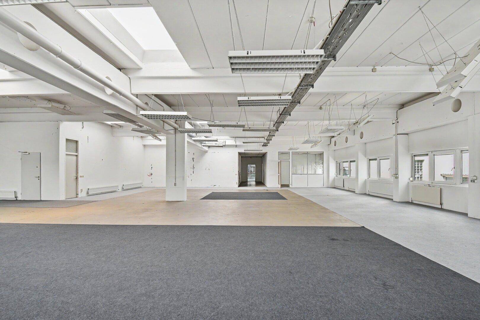 Weitläufiger, offener Bürobereich mit hohen Decken, industriellem Charme und unterschiedlichen Bodenbelägen.