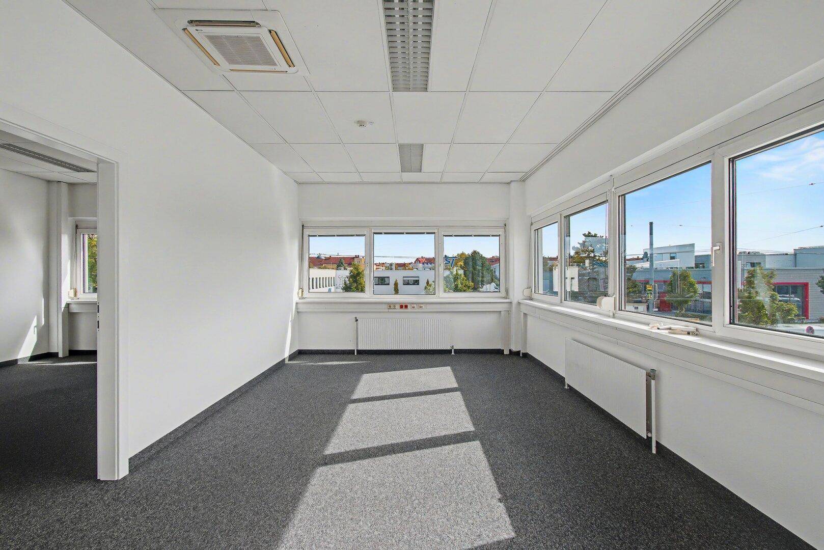 Helles Büro mit Teppichboden, großen Fenstern und Blick auf die umliegende Bebauung.