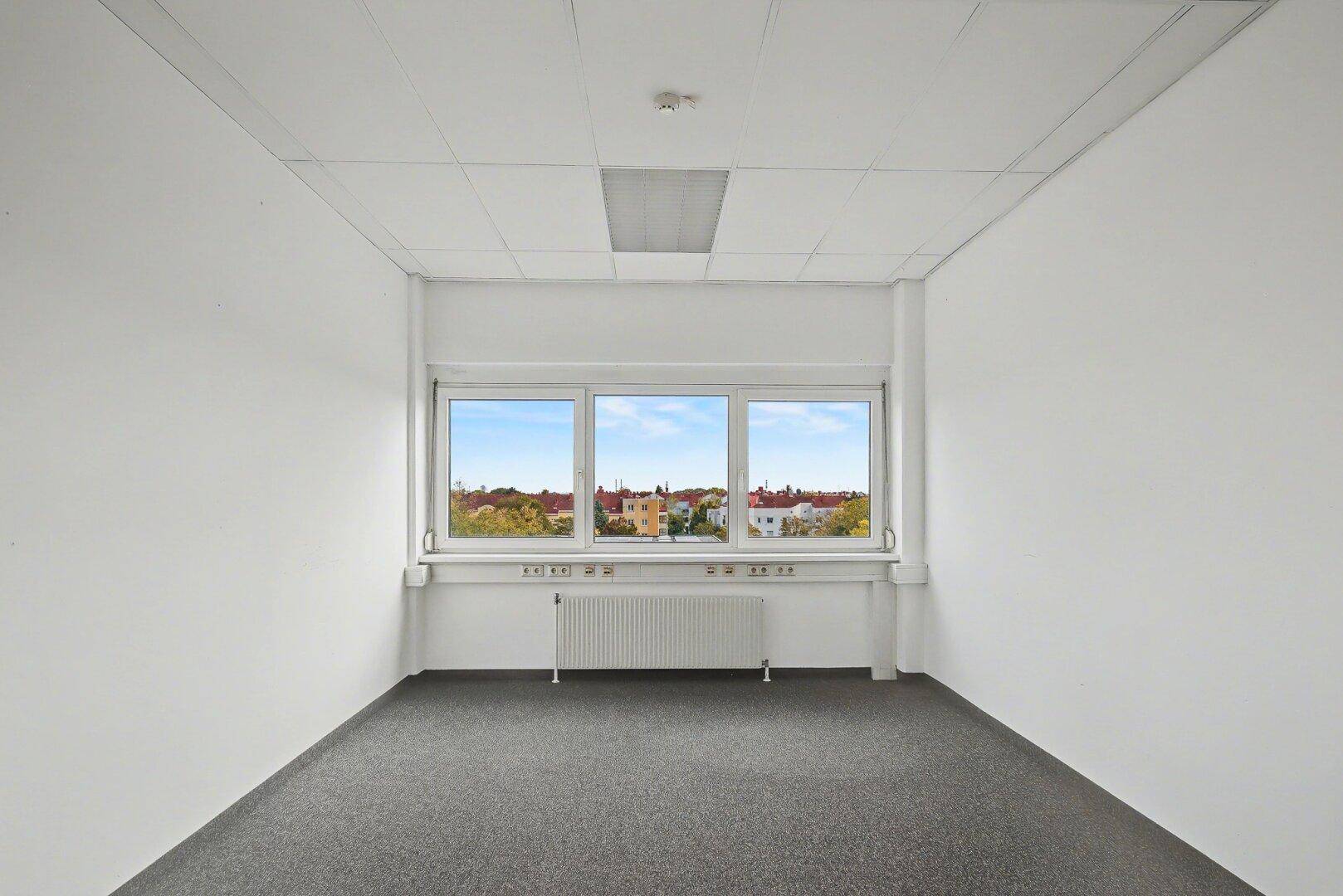 Leeres Büro mit grauem Teppichboden, weißen Wänden und einem Fenster mit Blick auf die Dächer.