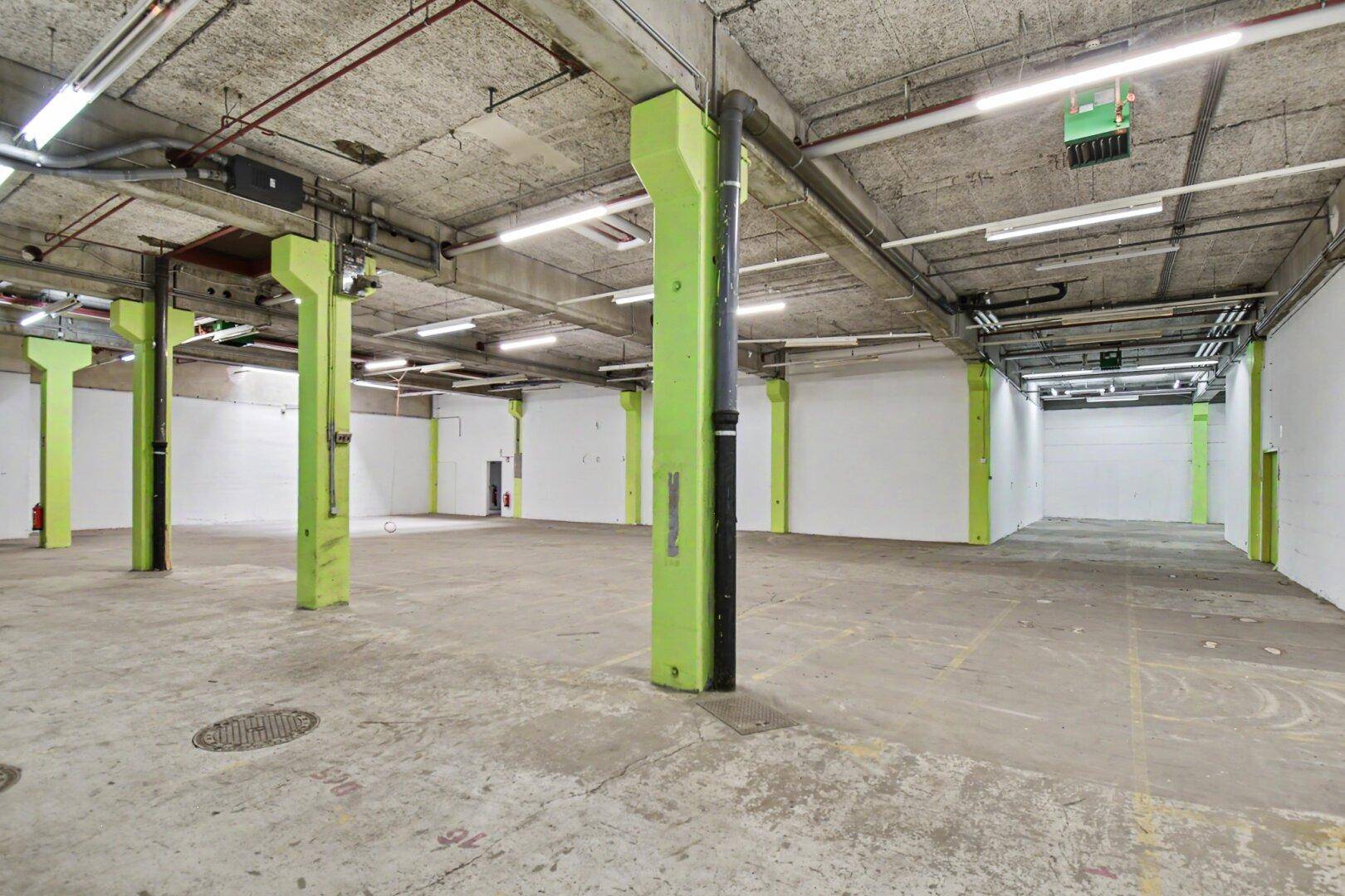 Geräumiger Industriebereich mit Betonboden, grünen Säulen und sichtbaren Deckeninstallationen.