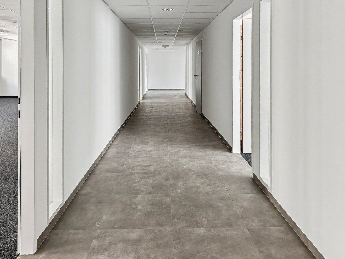 Langer, heller Flur mit grauen Fliesen, weißen Wänden und einer abgehängten Decke, der zu weiteren Räumen führt.