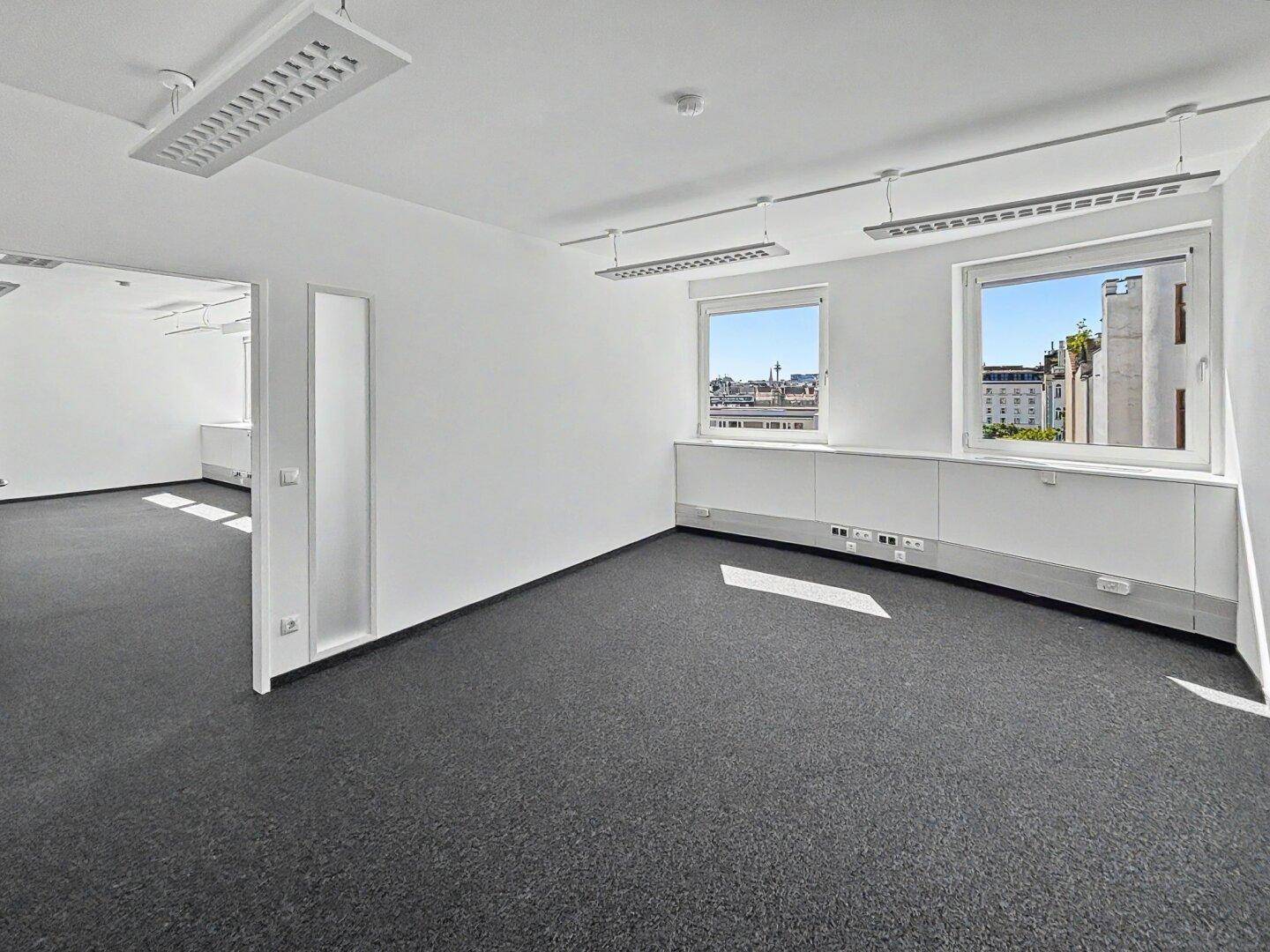 Heller Büroraum mit grauem Teppichboden, weißen Wänden und Fenstern, die einen Blick auf die Stadt bieten.