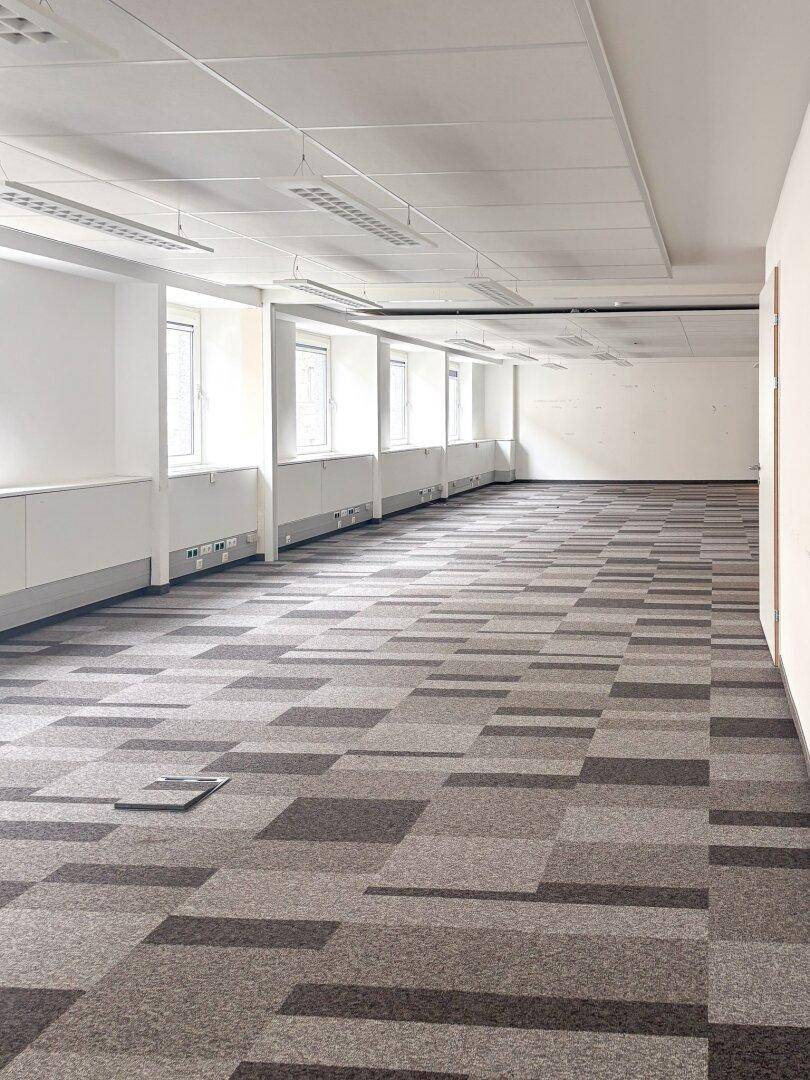 Langer, leerer Bürobereich mit gemustertem Teppichboden, weißen Wänden und vielen Fenstern für Tageslicht.