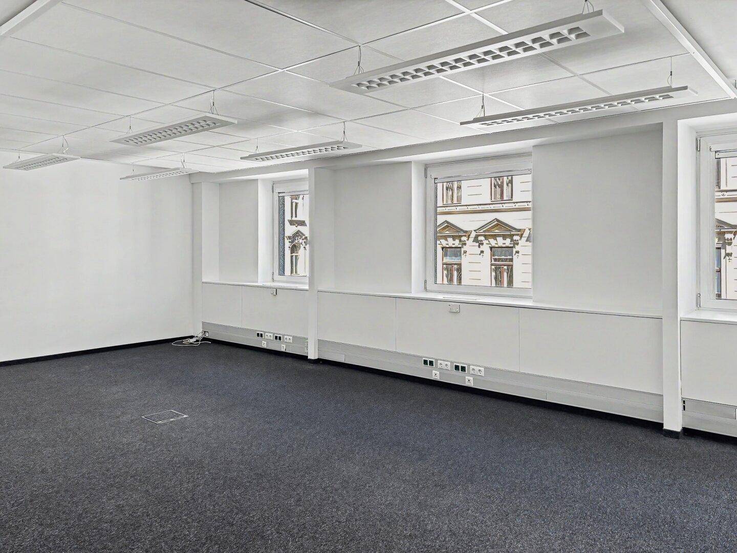 Großes, leeres Büro mit Teppichboden und mehreren Fenstern, die einen Blick auf die Umgebung bieten.