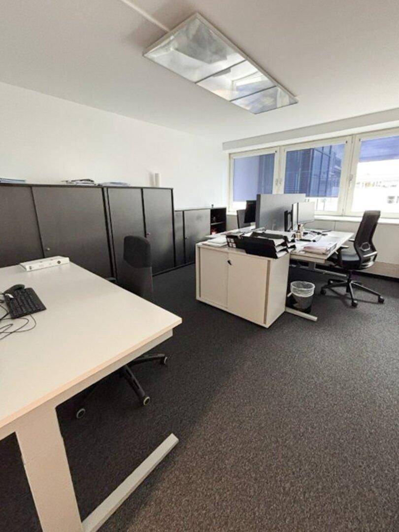 Möblierter Büroraum mit Arbeitsplätzen, schwarzen Schränken und Teppichboden, bereit zur Nutzung.