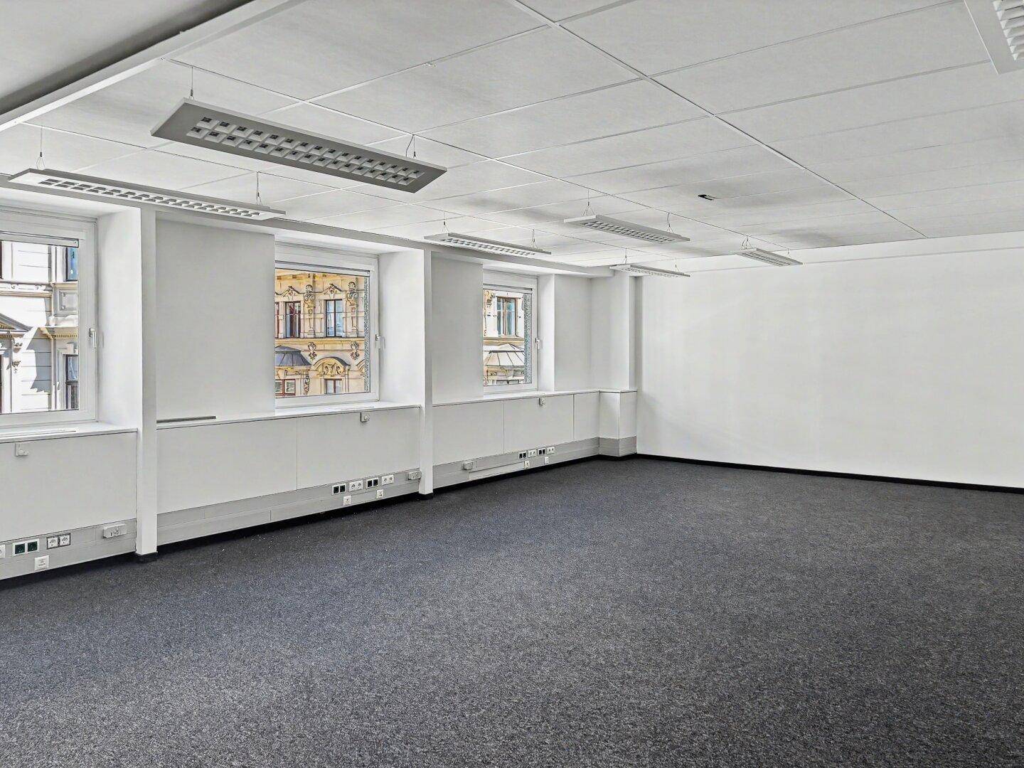 Leeres Büro mit grauem Teppichboden, weißen Wänden und mehreren Fenstern, die viel Tageslicht hereinlassen.