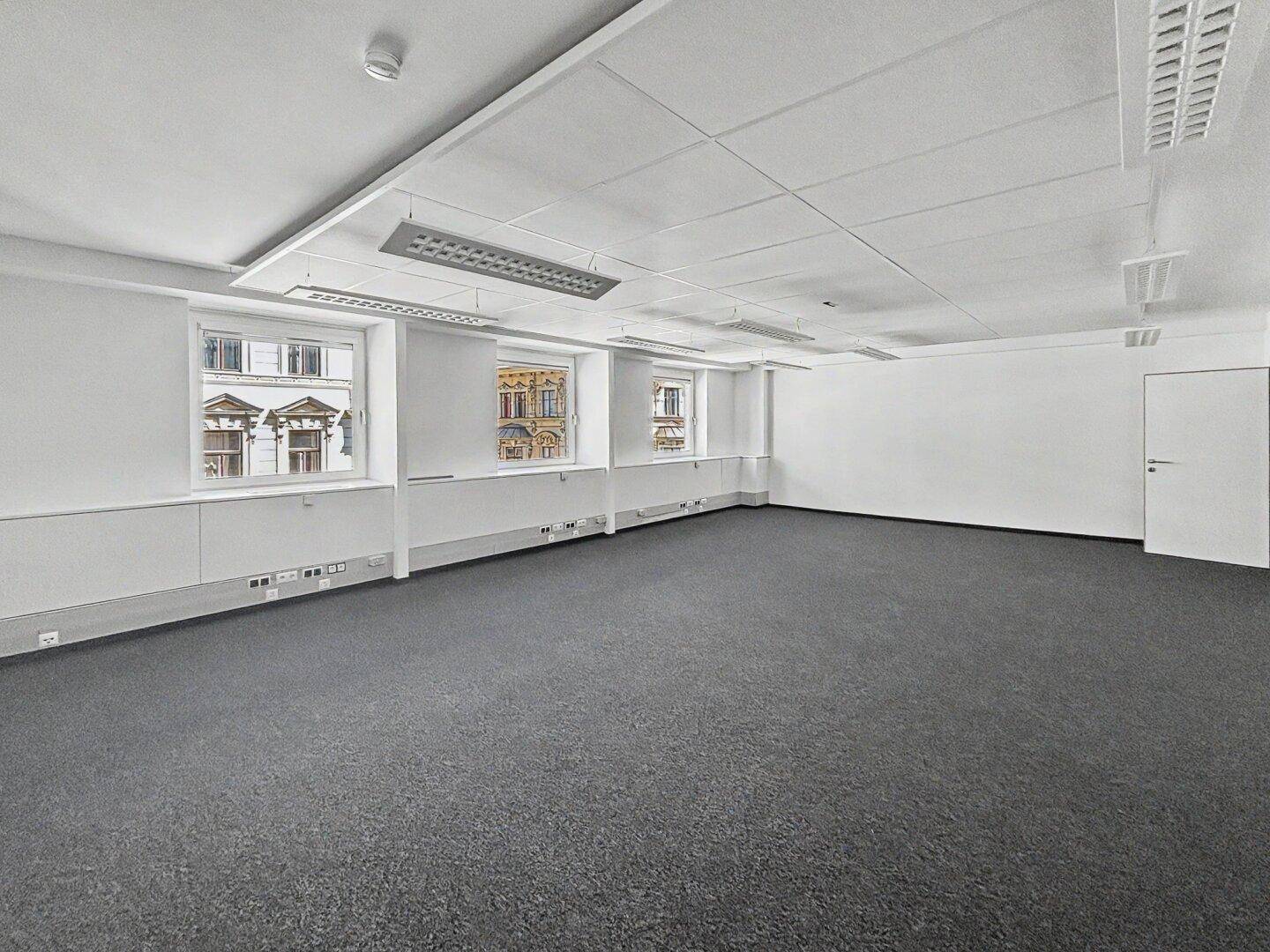 Geräumiges, unmöbliertes Büro mit grauem Teppichboden, weißen Wänden und einer modernen Tür.