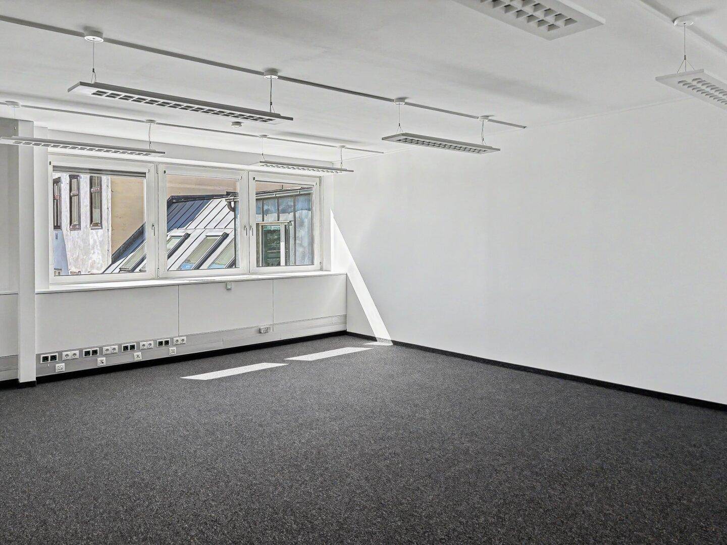 Helles, leeres Büro mit Teppichboden und großen Fenstern, die viel Sonnenlicht hereinlassen.