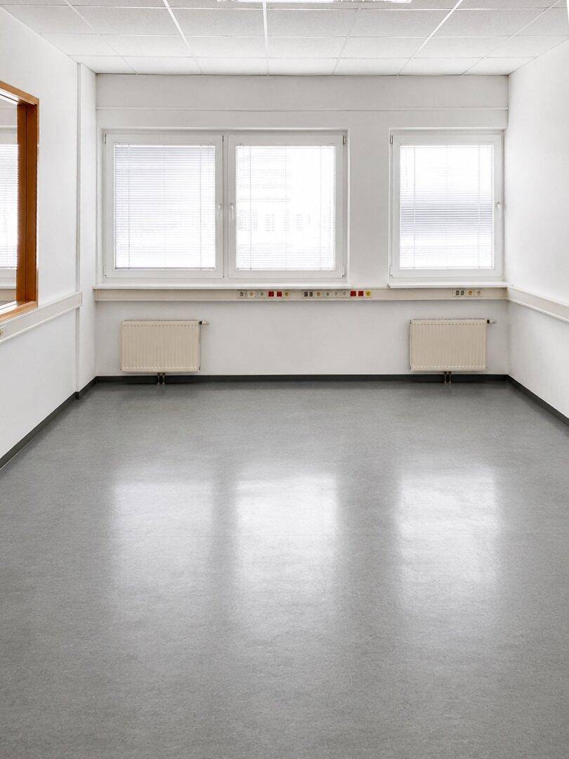Heller Büroraum mit drei großen Fenstern, Jalousien und einem gepflegten Vinylboden.