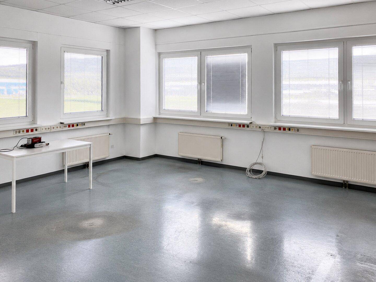 Geräumiges Büro mit mehreren Fenstern, Heizkörpern und einem einfachen Arbeitstisch.