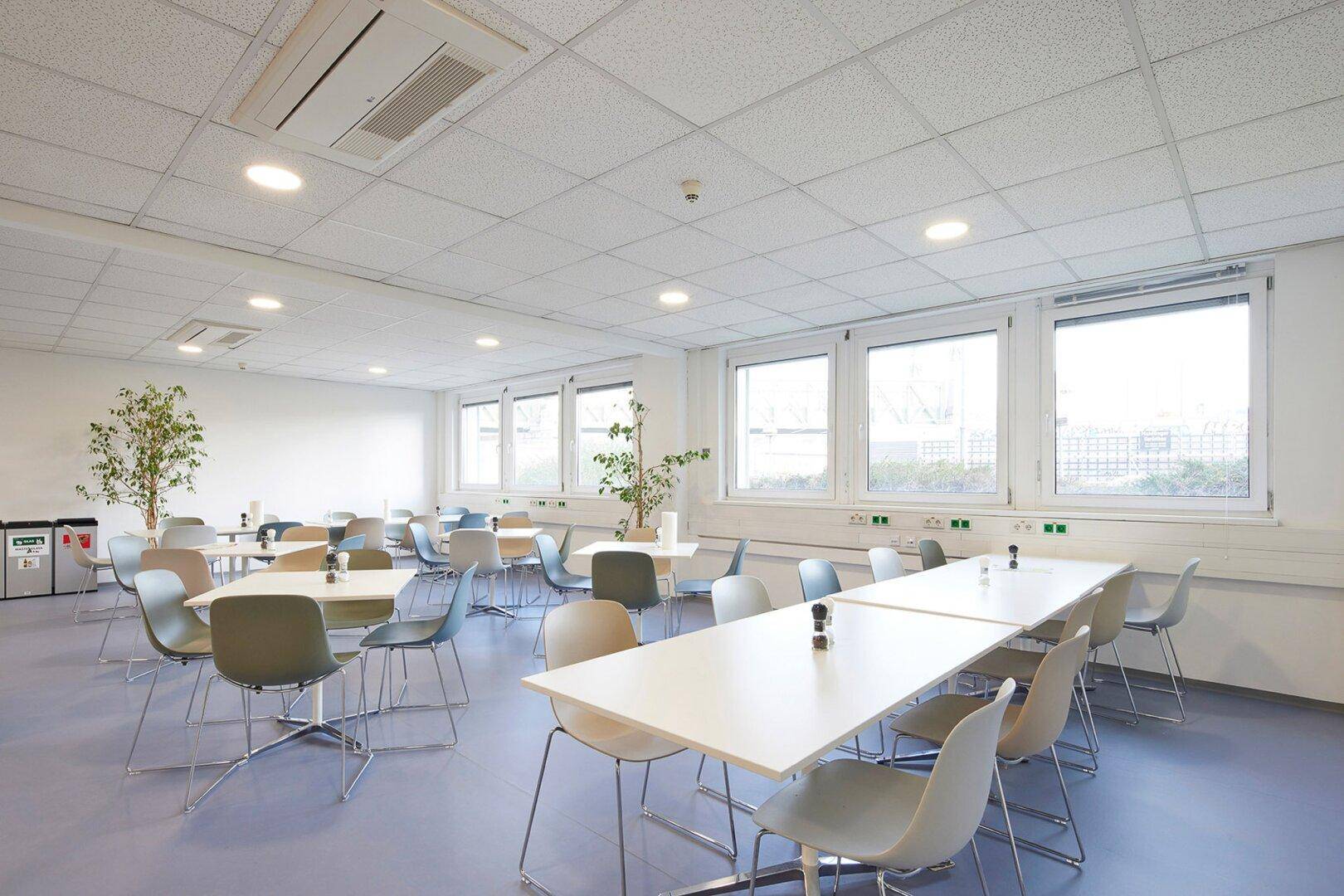 Geräumige Cafeteria mit vielen Tischen und Stühlen, großen Fenstern und hellem, modernem Ambiente.
