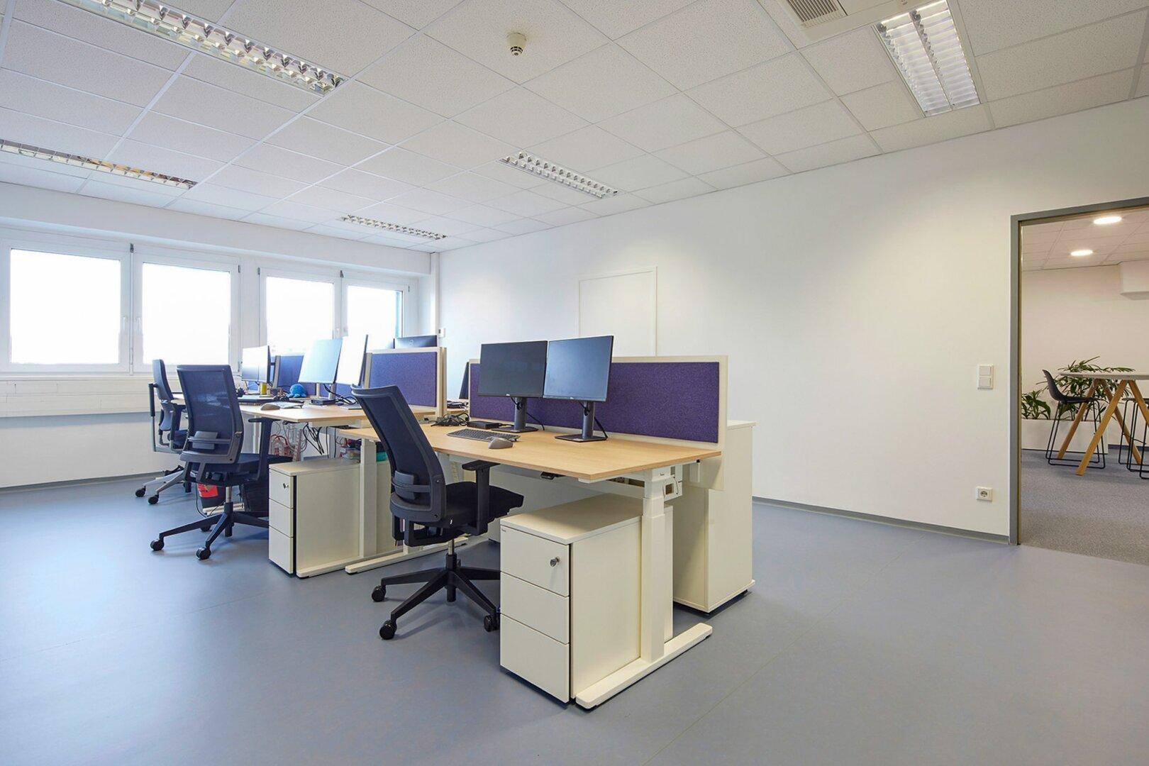 Offener Bürobereich mit mehreren Arbeitsplätzen, ergonomischen Stühlen und großen Fenstern für viel Tageslicht.