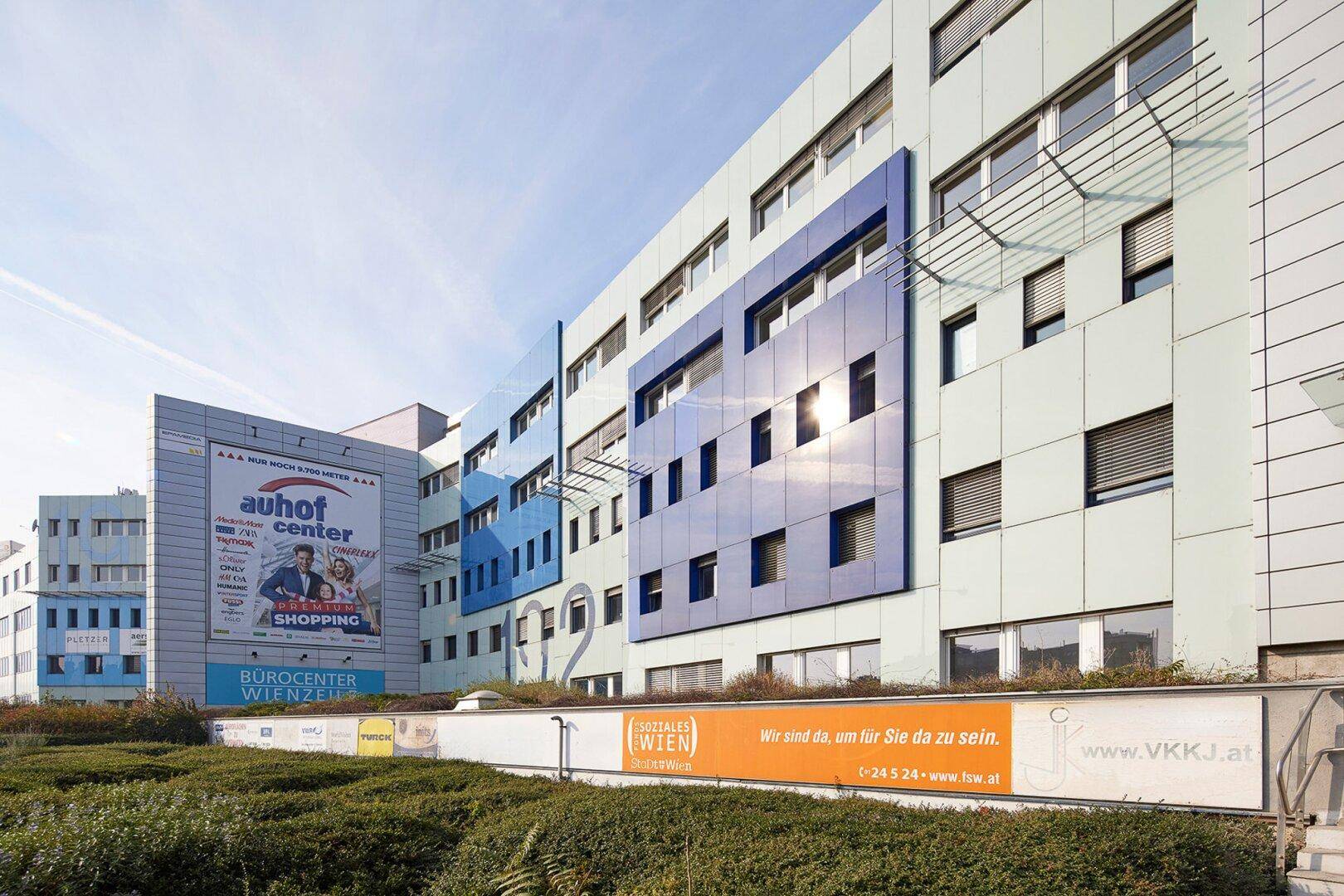 Modernes Bürogebäude mit blauer und weißer Fassade unter klarem Himmel, umgeben von Grünflächen.