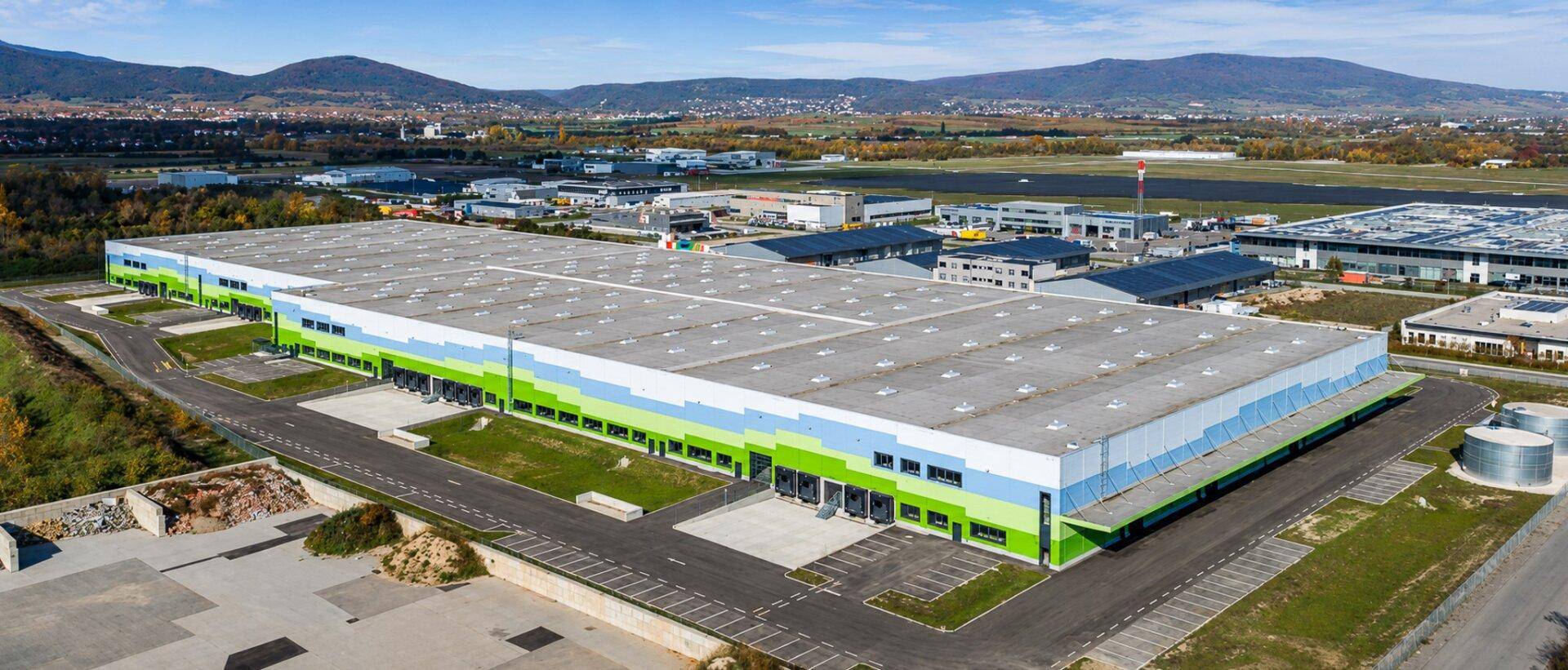 Großes modernes Industriegebäude mit grüner und blauer Fassade, umgeben von weiteren Gewerbebauten und Bergen im Hintergrund.
