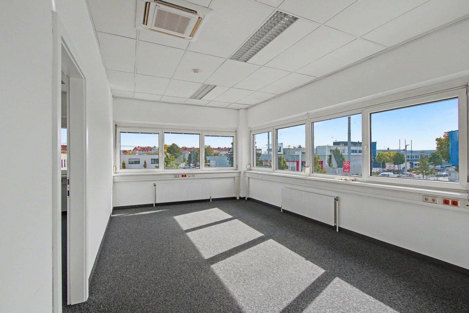 Helles Büro mit Teppichboden, großen Fenstern und Blick auf die umliegende Bebauung.