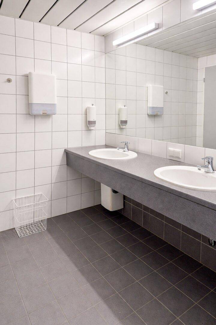 Öffentlicher Waschraum mit zwei Waschbecken, großem Spiegel und weißen Fliesen an Wänden und Boden.