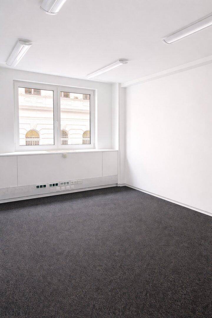 Heller, leerer Büroraum mit Teppichboden und großen Fenstern, die Tageslicht hereinlassen.