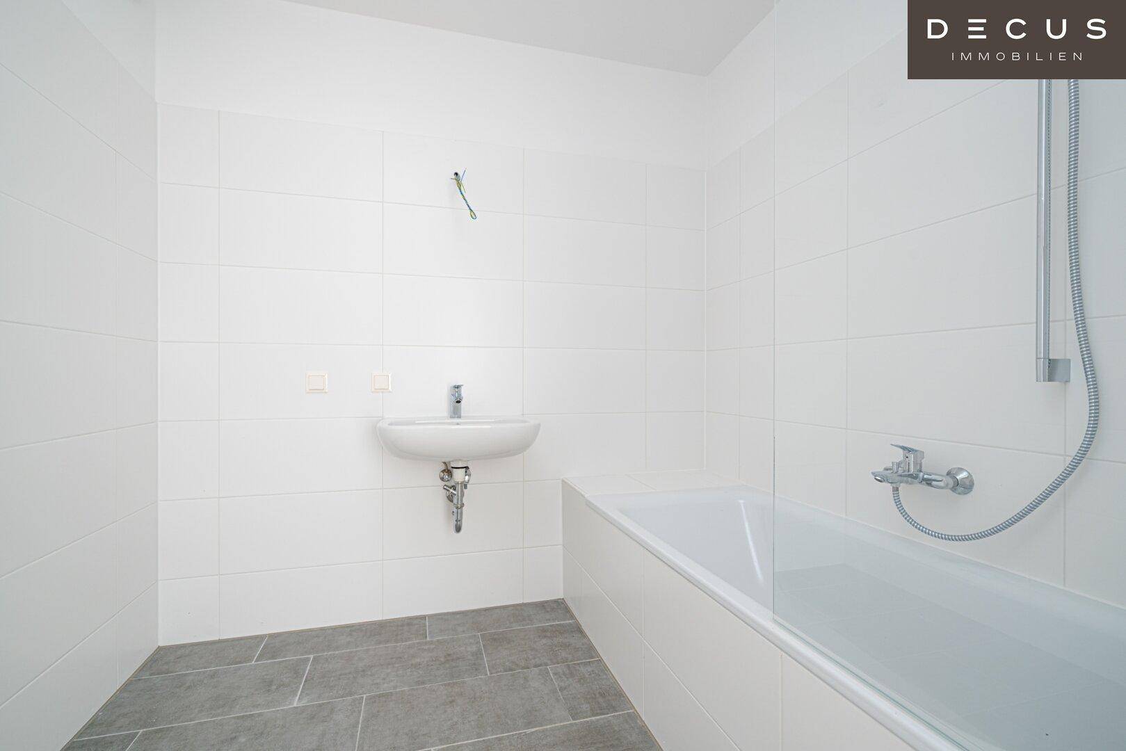 Unmöbliertes Badezimmer mit weißen Wandfliesen, grauen Bodenfliesen, Waschbecken und Badewanne mit Duschfunktion.