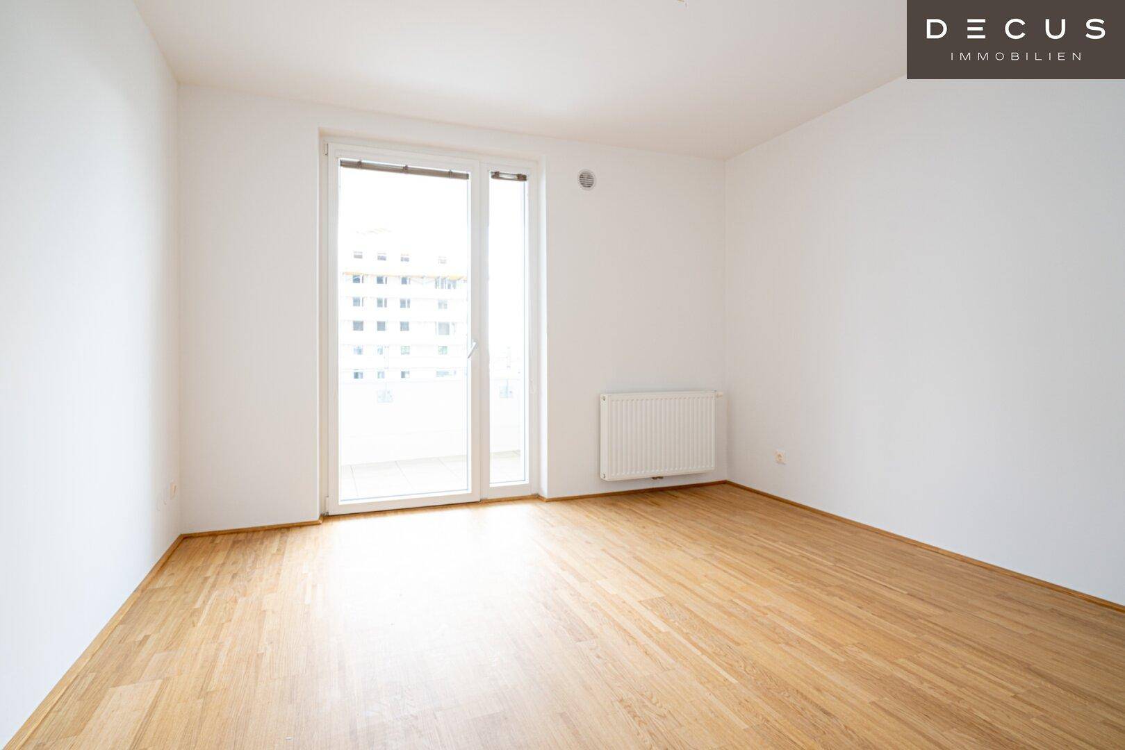 Helles, leeres Zimmer mit Holzboden und Zugang zu einem Balkon oder einer Terrasse.