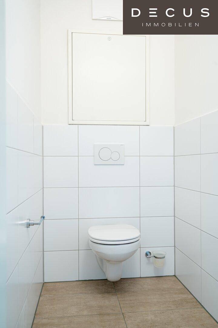 Separates WC mit wandhängender Toilette und weißen Fliesen an den Wänden und auf dem Boden.
