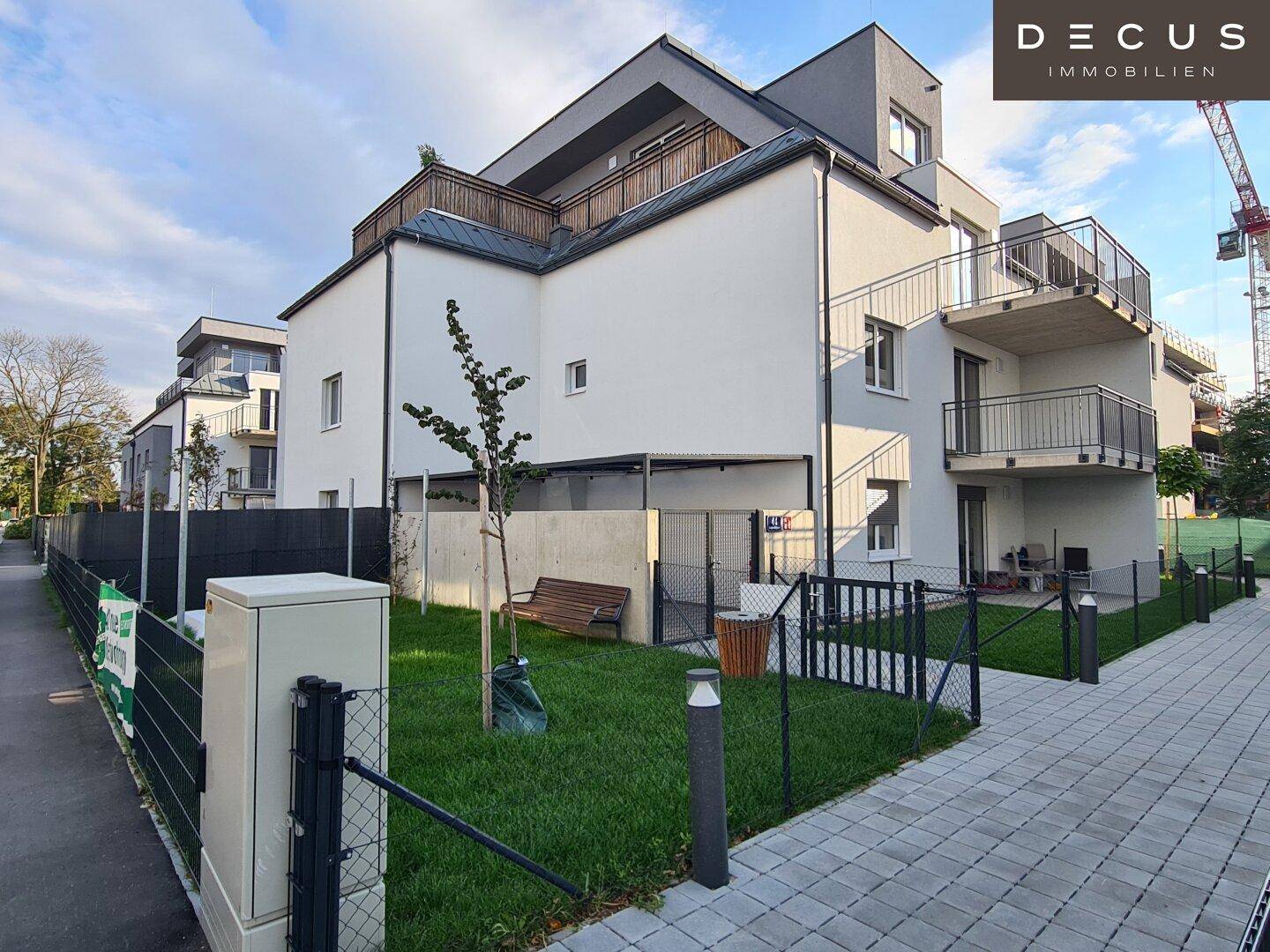 Ansicht des modernen Apartmentgebäudes mit Balkonen, kleiner Grünfläche und einer Sitzbank im Freien.