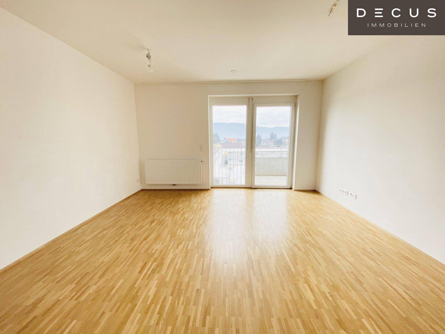 Geräumiges, leeres Zimmer mit Holzboden und großen Fenstern, die viel Tageslicht hereinlassen.