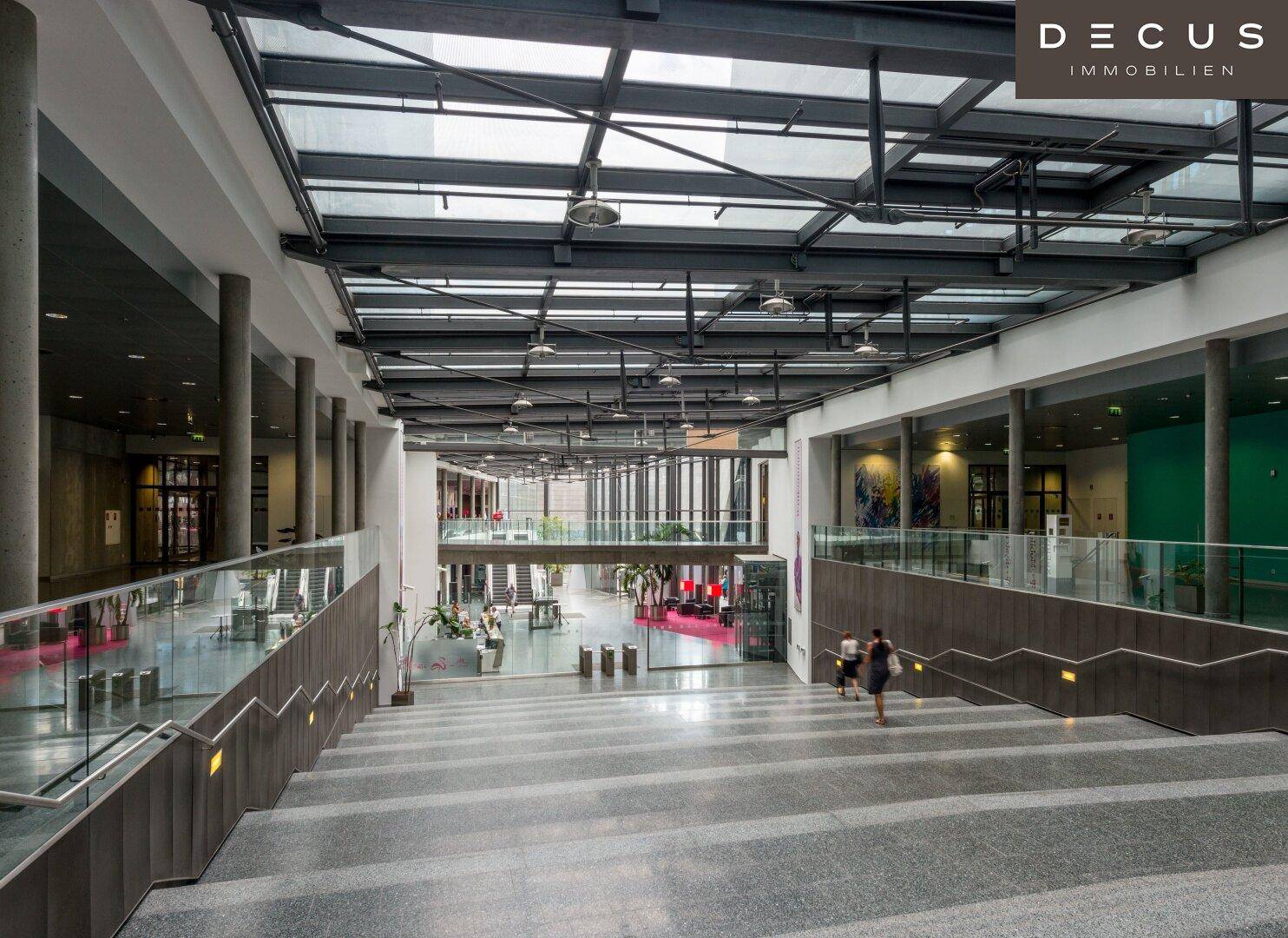 Geräumige, moderne Lobby mit breiter Treppe, Glasgeländern und viel Tageslicht durch das Glasdach.