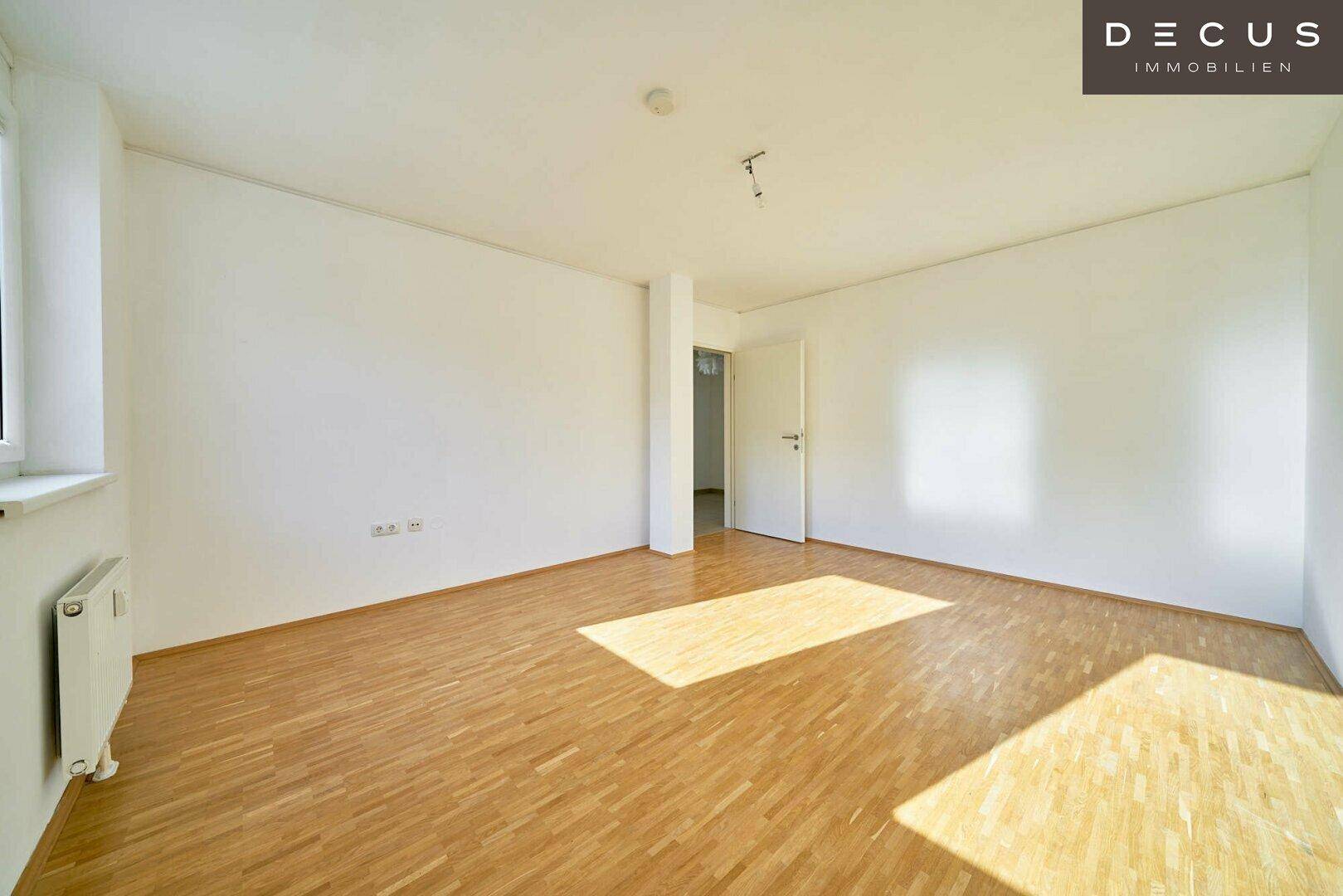 Helles, leeres Zimmer mit Parkettboden, weißen Wänden und viel natürlichem Licht.