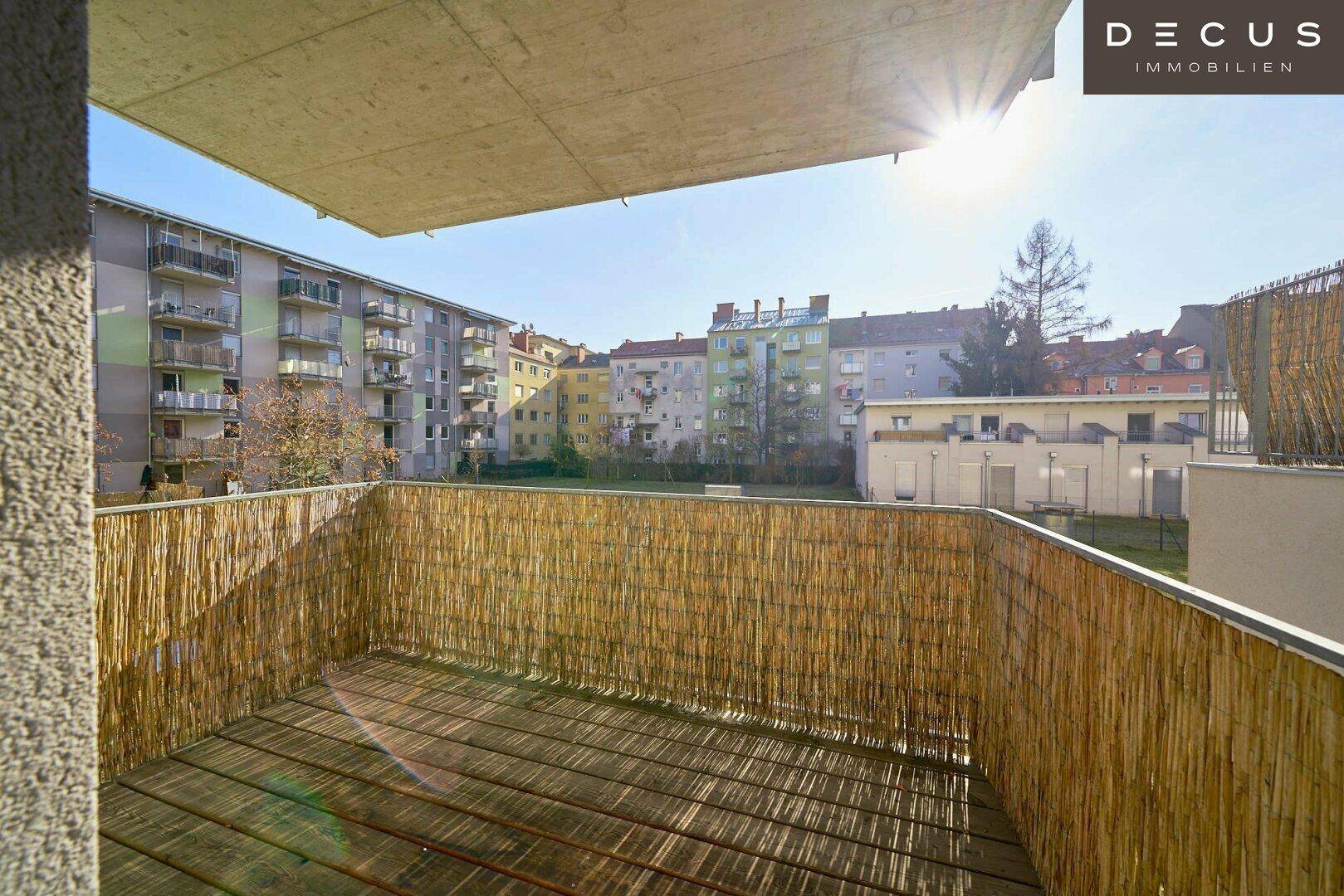 Sonniger Balkon mit Holzboden und natürlichem Bambus-Sichtschutz, umgeben von Wohngebäuden.