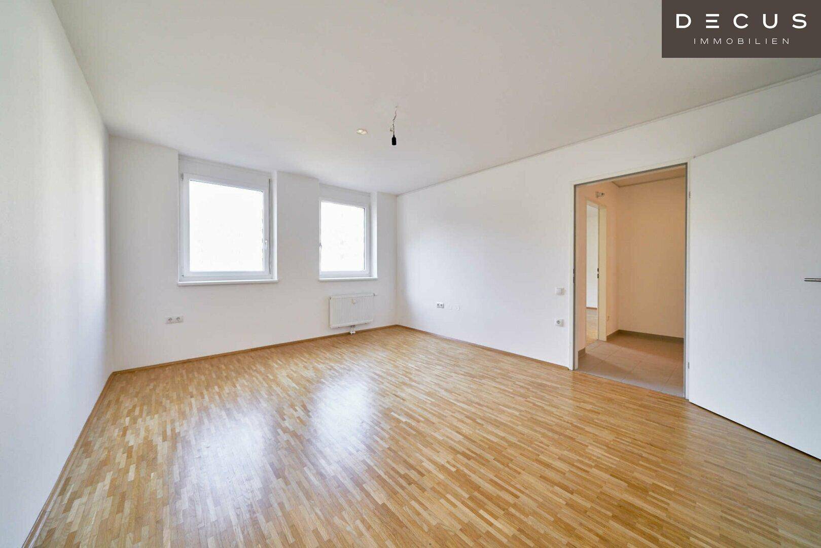 Heller Raum mit Parkettboden und zwei Fenstern, bietet Zugang zu einem weiteren Bereich.