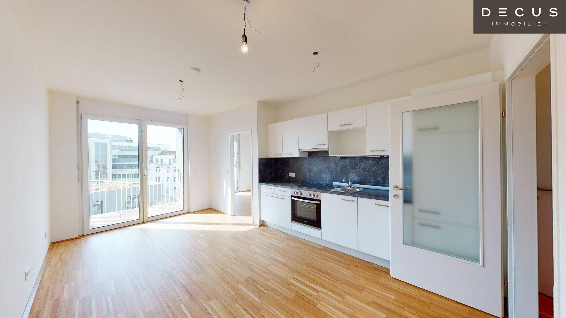 Heller offener Wohnbereich mit integrierter Küche, großen Fenstern und Zugang zu einem Balkon.
