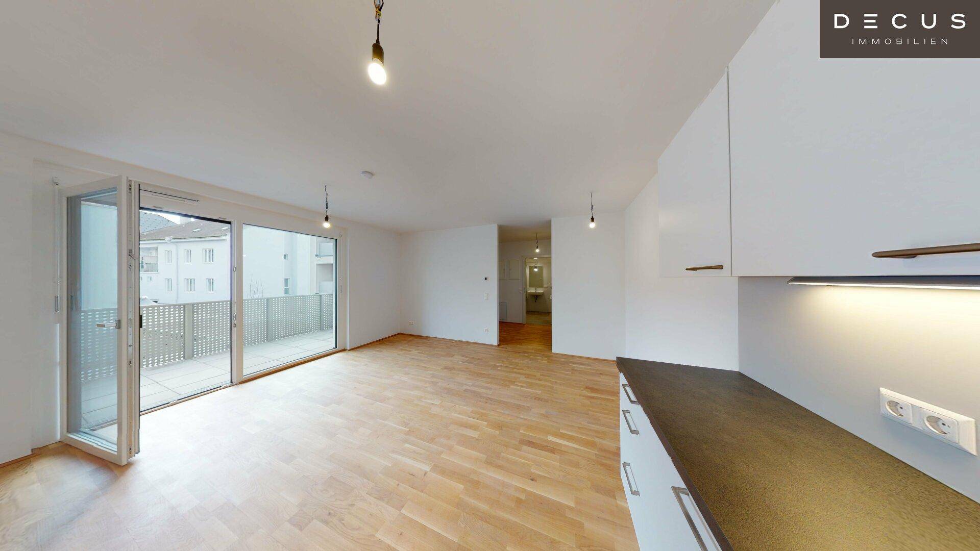 Heller offener Wohnbereich mit Holzboden, großen Schiebetüren zum Balkon und integrierter Küchenzeile.