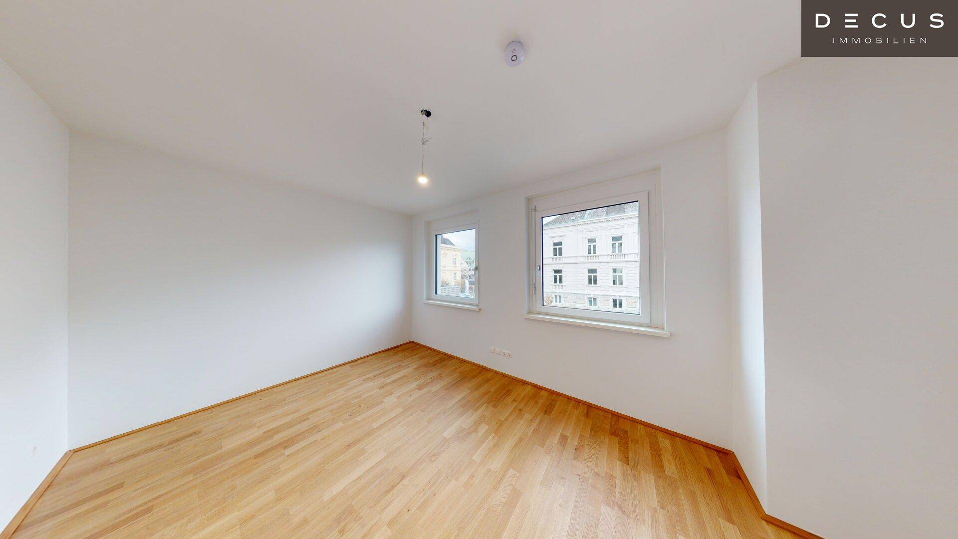 Helles Schlafzimmer mit Parkettboden und zwei Fenstern, die viel Tageslicht hereinlassen.