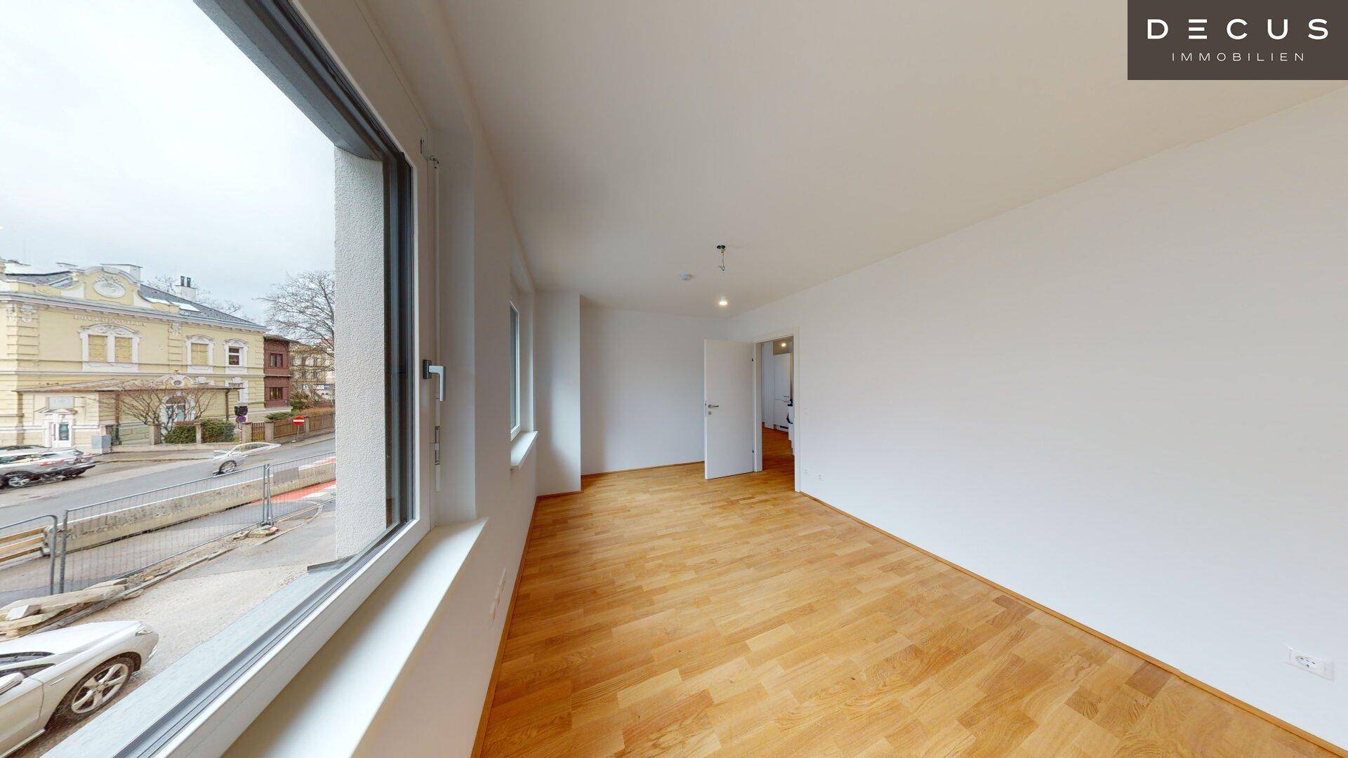 Geräumiges Wohnzimmer mit Parkettboden und großen Fenstern mit Blick auf die Straße.