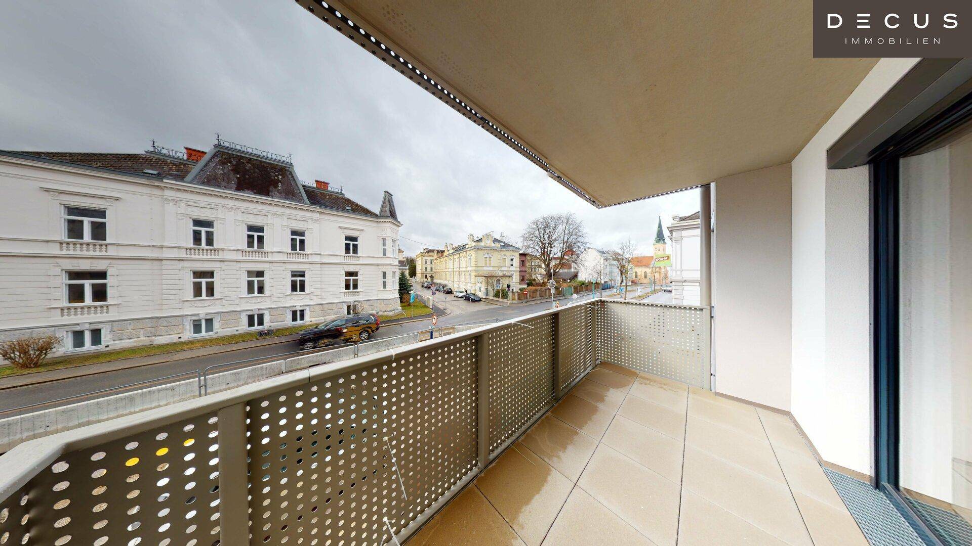 Heller Balkon mit Fliesenboden und Blick auf eine belebte Straße und historische Gebäude.