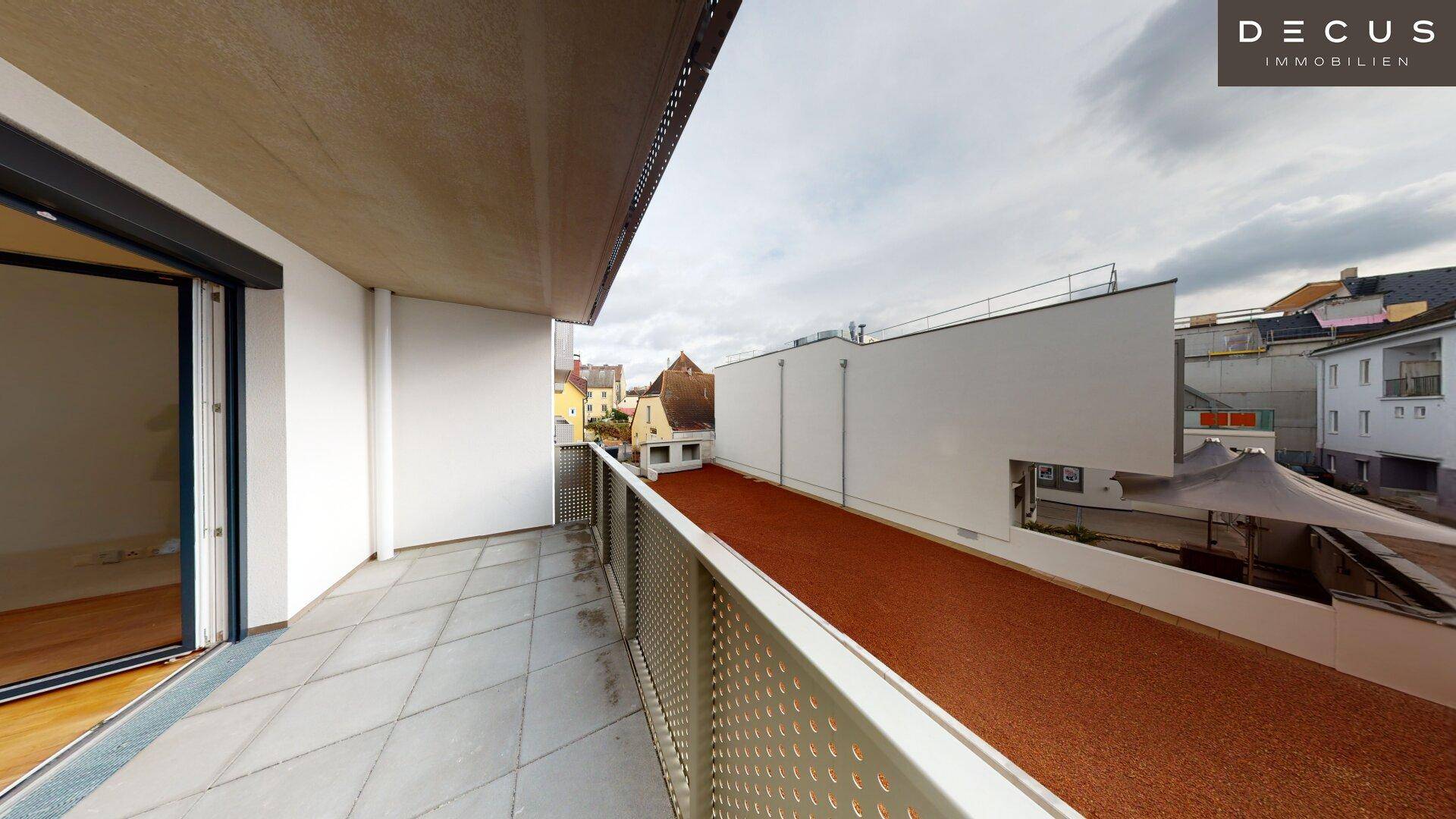 Langer Balkon mit Fliesenboden und Blick auf eine begrünte Fläche und Nachbargebäude.