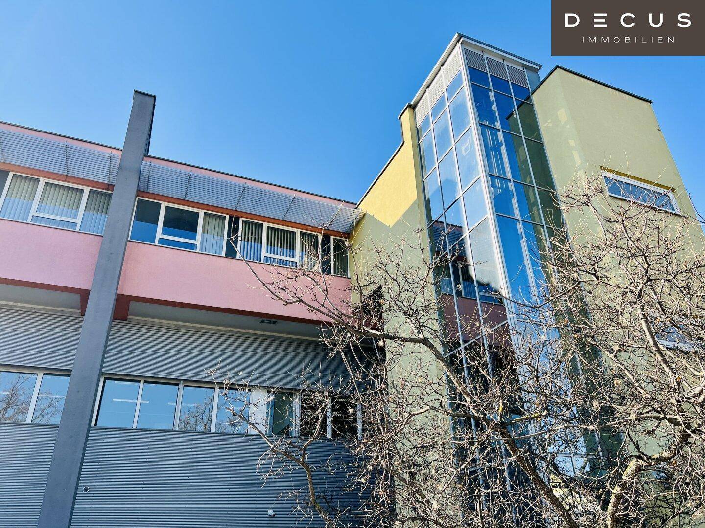 Ansicht eines modernen Bürogebäudes mit Glasaufzug und farbigen Fassadenelementen unter klarem Himmel.