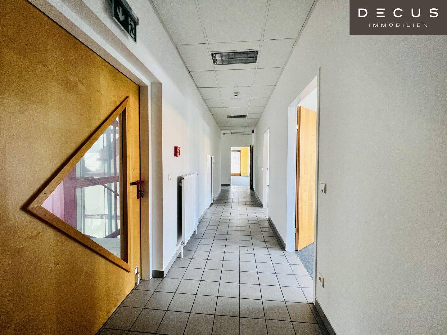 Langer Flur mit Fliesenboden, weißen Wänden und Deckenbeleuchtung, verbindet verschiedene Bereiche.